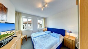 Ein Hotelzimmer mit zwei Betten, einem Schreibtisch mit Fernseher, einem Sessel, einer Lampe und einem Fenster mit Blick auf Gebäude.