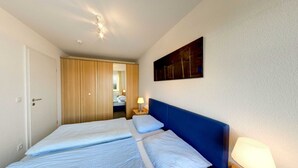 A hotel room with a double bed, a wooden wardrobe, a painting on the wall, and a ceiling light. - Villa Aquamarina Whg. 22, Fahrstuhl, Seeblick - Villa Aquamarina Whg. 22 (Heringsdorf)