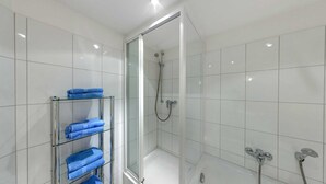 A modern bathroom with white tiles, a glass shower enclosure, and a glass shelf with folded towels. - Villa Aquamarina Whg. 22, Fahrstuhl, Seeblick - Villa Aquamarina Whg. 22 (Heringsdorf)