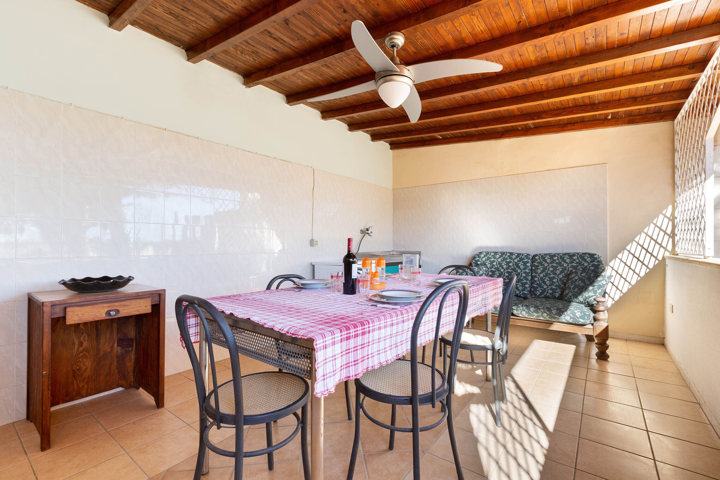 Un comedor con techo de madera, ventilador de techo, mesa con mantel a cuadros, sillas, sofá y una mesita con un bol y botellas.