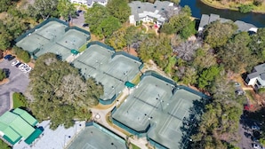 Vista aérea de un complejo deportivo con varias canchas de tenis, rodeado de árboles y edificios residenciales.