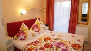A bedroom with a red headboard, floral bedding, a bedside lamp, and a window with red curtains.