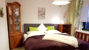 Un lit bien fait, avec une tête de lit de couleur foncée, une armoire en bois aux portes vitrées et une petite table de chevet avec une lampe.