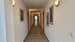 Hotellin käytävä, jossa on puulattia, seinillä on kehystettyjä taideteoksia ja seinään kiinnitetyt valaisimet.