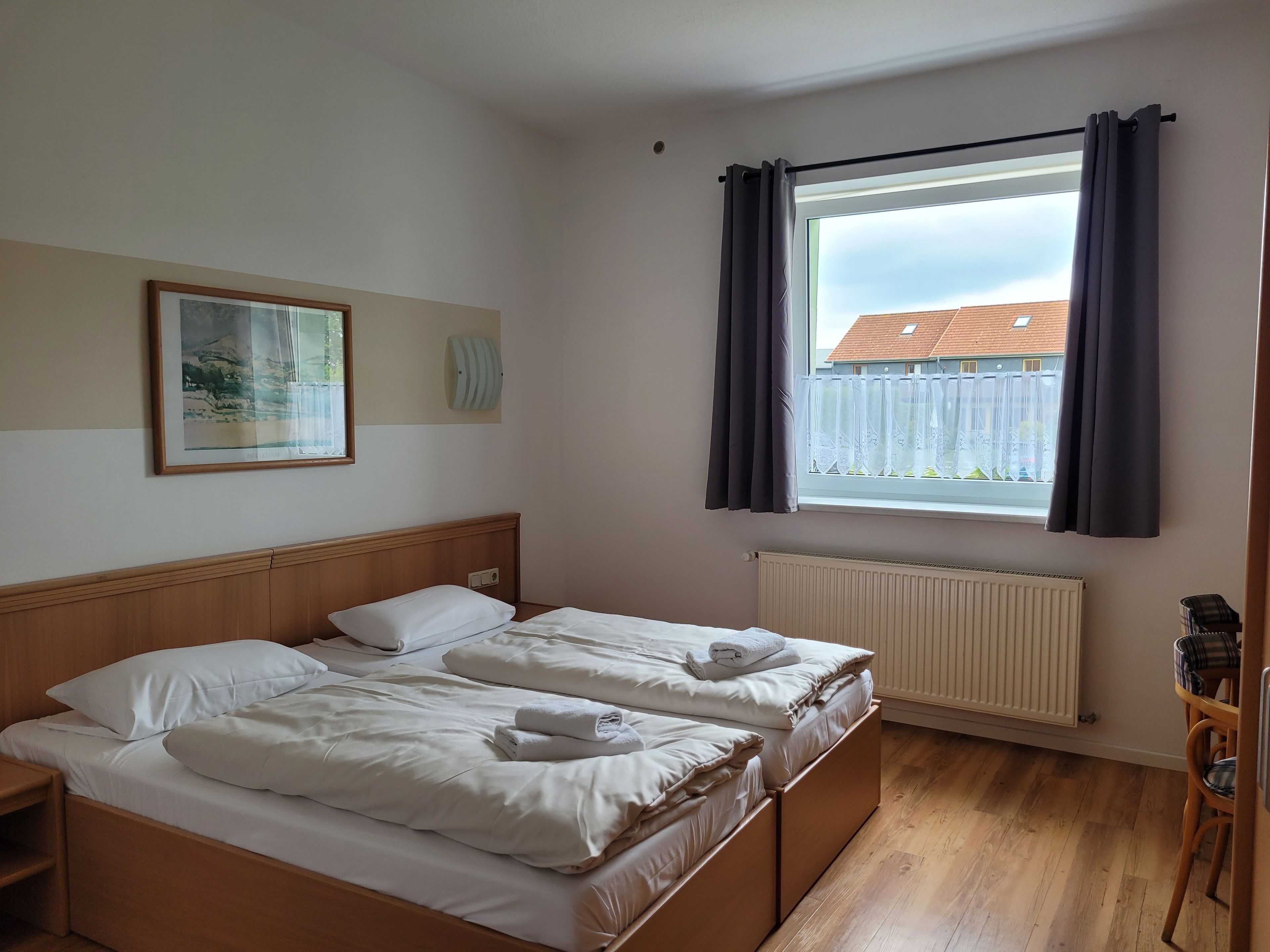 Ein Schlafzimmer mit zwei Betten, einem Fenster mit Vorhängen, einem Heizkörper, einem Holzboden und einem gerahmten Bild an der Wand.