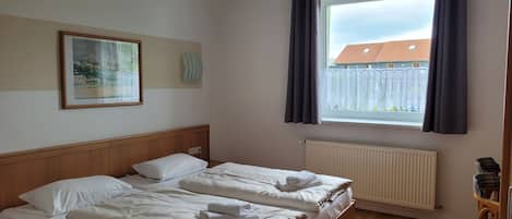 A bedroom with two beds, a window with curtains, a radiator, a wooden floor, and a framed picture on the wall.