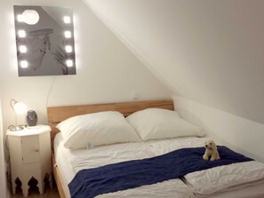 A bedroom with a wooden headboard, white bedding, a small bedside table with a lamp, and a wall-mounted mirror with lights.