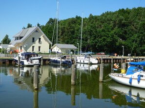 Ein Yachthafen mit mehreren vertäuten Booten, ein Haus mit geneigtem Dach und ein bewaldeter Hügel im Hintergrund.