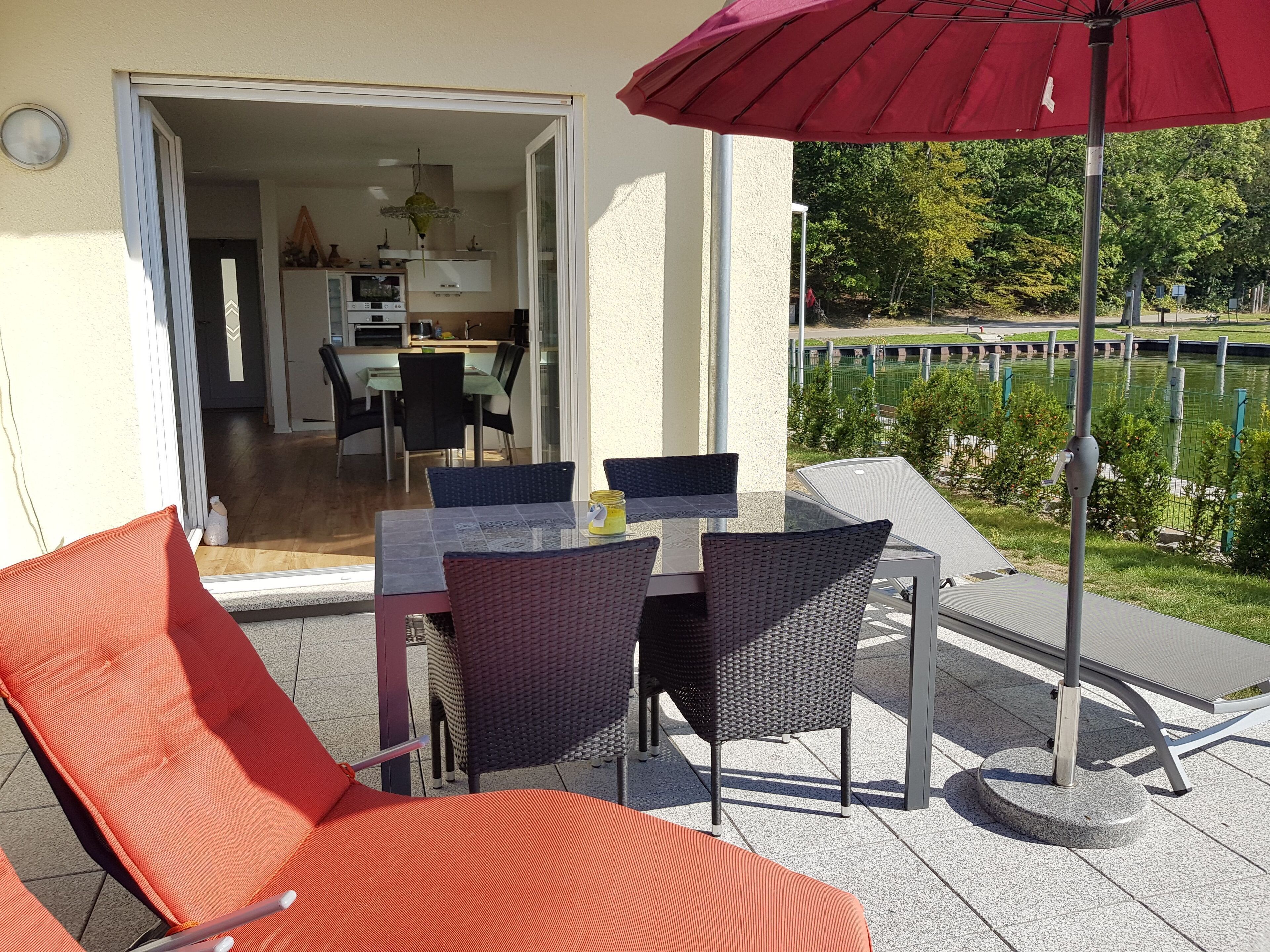 Eine Terrasse im Freien mit Tisch und Stühlen, einem roten Sonnenschirm und Blick auf einen Garten sowie ein Gebäude im Hintergrund.