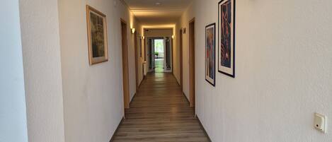 A hotel hallway with wooden flooring, framed artwork on the walls, and wall-mounted lighting.