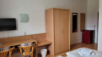 Una habitación con un escritorio de madera, dos sillas, un televisor, un armario, una cama con toallas, un cubo de basura y un refrigerador rojo.