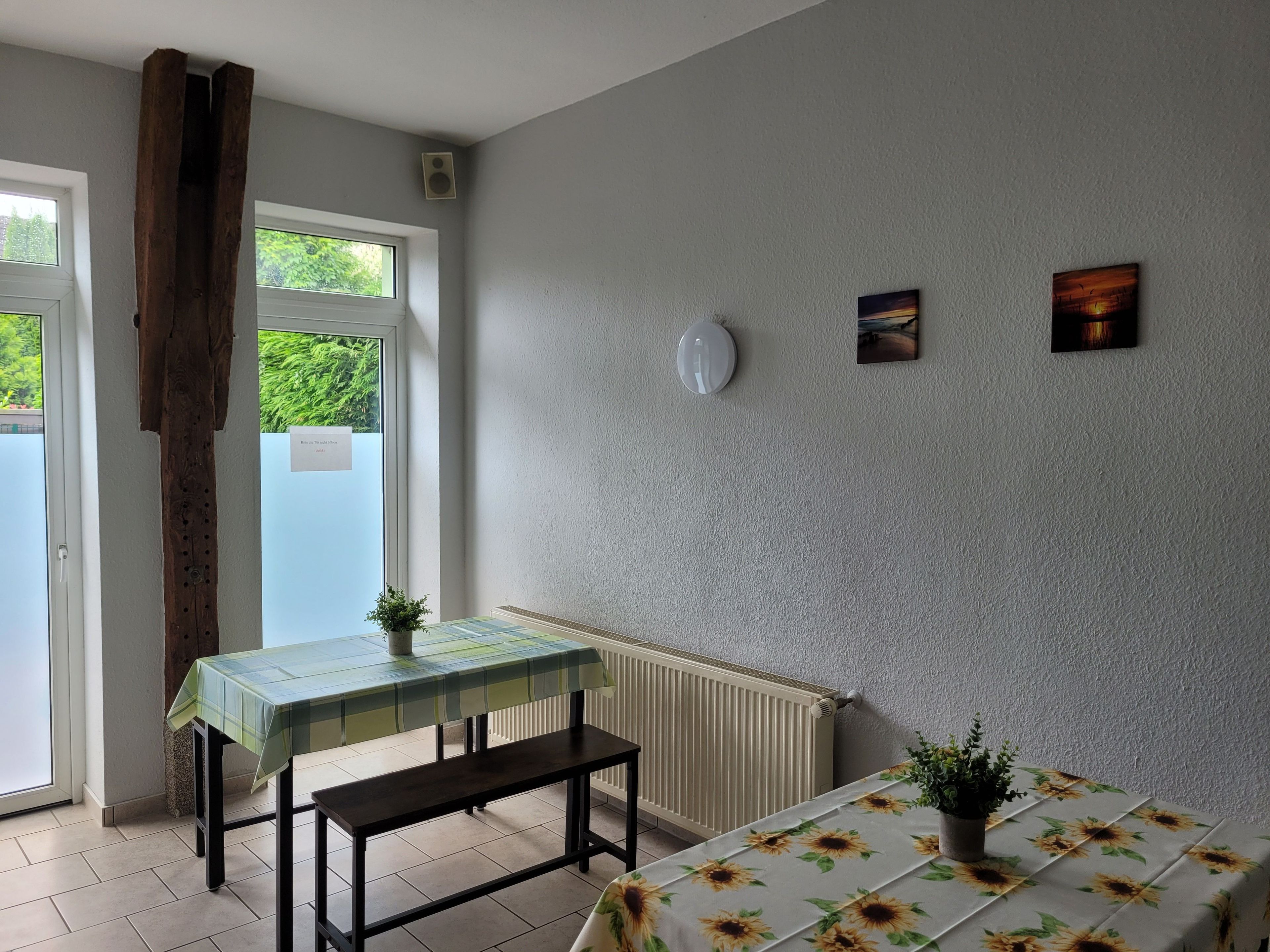 Une pièce comprenant une table, un banc, deux plantes en pot, un radiateur et des œuvres d’art accrochées au mur.