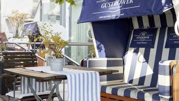 An outdoor seating area with striped cushions and a table, a potted plant, and a blue awning with the text "GÜSTROWER".
