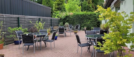 A patio area with tables and chairs, surrounded by potted plants and a brick floor.