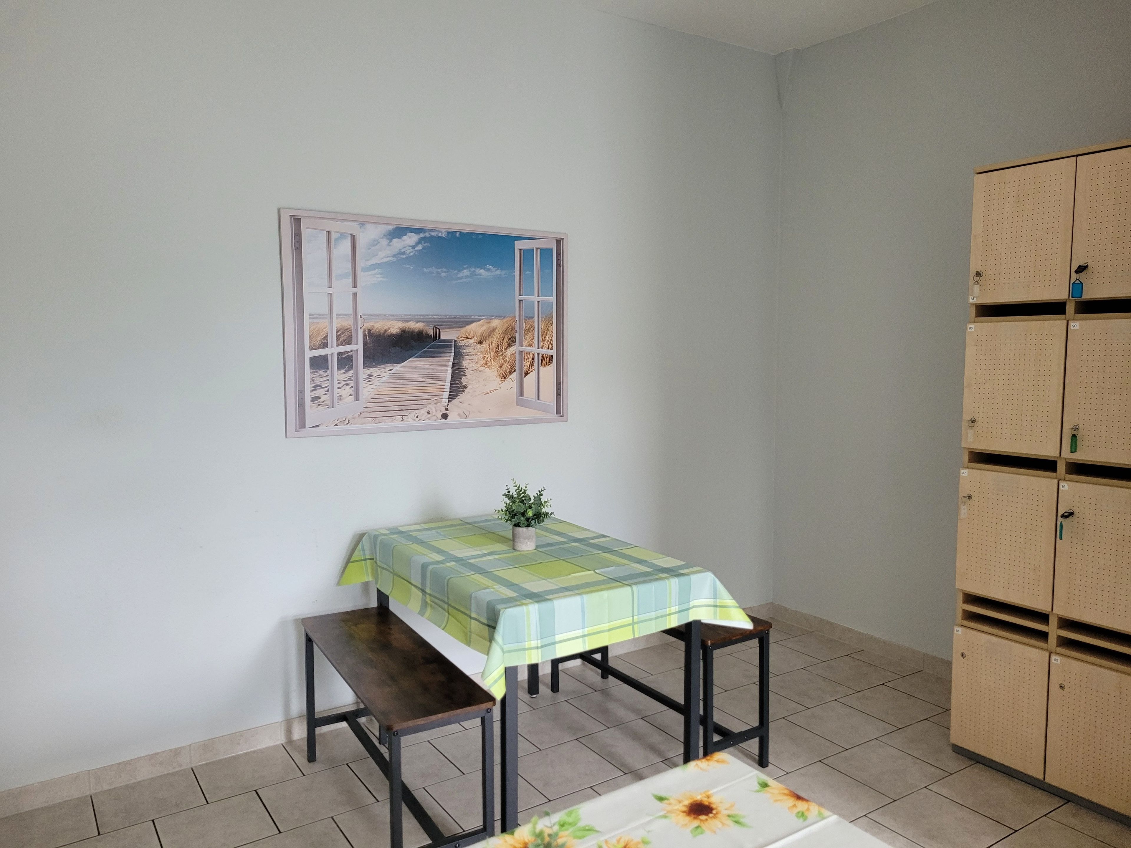 Um quarto com uma mesa e bancos, um armário com vários compartimentos e um quadro na parede retratando uma cena de praia.