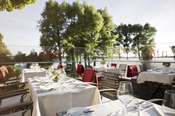 Un espace de restauration en plein air avec des tables prévues pour deux personnes, entouré d’arbres et d’une rambarde.