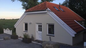 Ein kleines, eingeschossiges Haus mit rotem Ziegeldach, weißen Außenwänden und einer weißen Tür. Auf der Vorderseite befinden sich zwei Fenster, eines mit einem kleinen Vordach. Das Haus wird von einem Schotterweg und einem weißen Zaun umgeben.