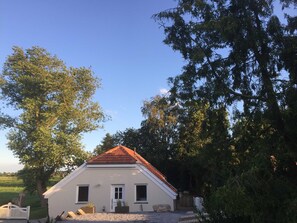 A small, white building with a red roof, surrounded by trees and a gravel path.