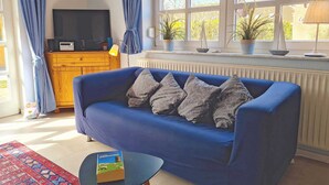 Una sala de estar con un sofá azul, una mesita con un libro, un televisor y una ventana con cortinas.