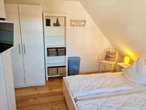 Une chambre à coucher comprenant un lit, une armoire, une étagère avec des paniers, une chaise et une poubelle.