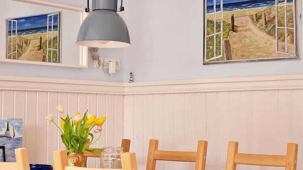 Un comedor con una mesa de madera, sillas, un florero con flores, una lámpara y dos cuadros enmarcados con escenas costeras.