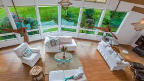 Ein geräumiges Wohnzimmer mit großem Fenster und Blick auf einen üppigen Garten, mit einer weißen Sofagarnitur, einem kleinen runden Tisch und einem Deckenventilator.