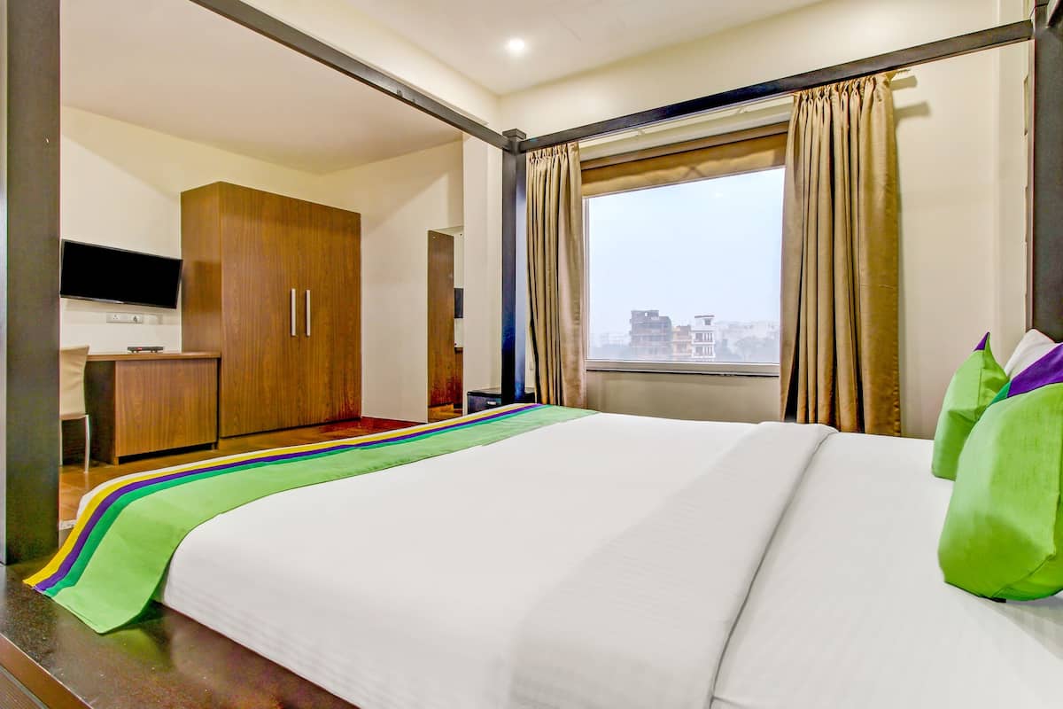 Habitación de hotel con cama, televisor sobre un soporte, armario y ventana con cortinas.