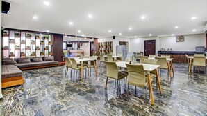 Une vaste salle à manger d’hôtel, dotée d’un sol en marbre, de nombreuses tables et chaises, d’un canapé et d’un coin cuisine à l’arrière-plan.