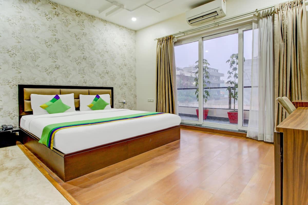 Habitación de hotel con cama, escritorio de madera, ventana con cortinas y balcón con plantas en macetas.