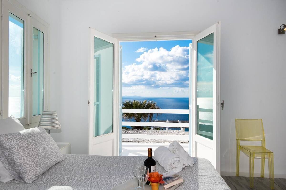 A bedroom with a bed, a nightstand, a lamp, a chair, and a view of the sea through open doors.