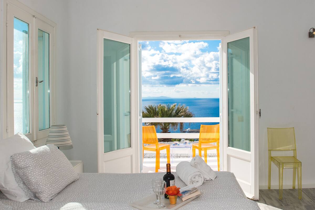 A room with a bed, a nightstand, a lamp, and a view of the sea through open doors.