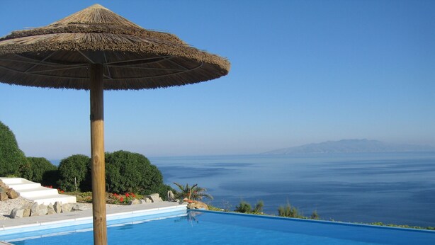 En swimmingpool med parasol af strå, omgivet af en anlagt have med trapper og beplantning, med udsigt over et vandareal.