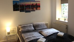 A bedroom with a bed, pillows, a nightstand, a wall-mounted artwork, a window, and a radiator. - Robs Ferienwohnung (Dresden)
