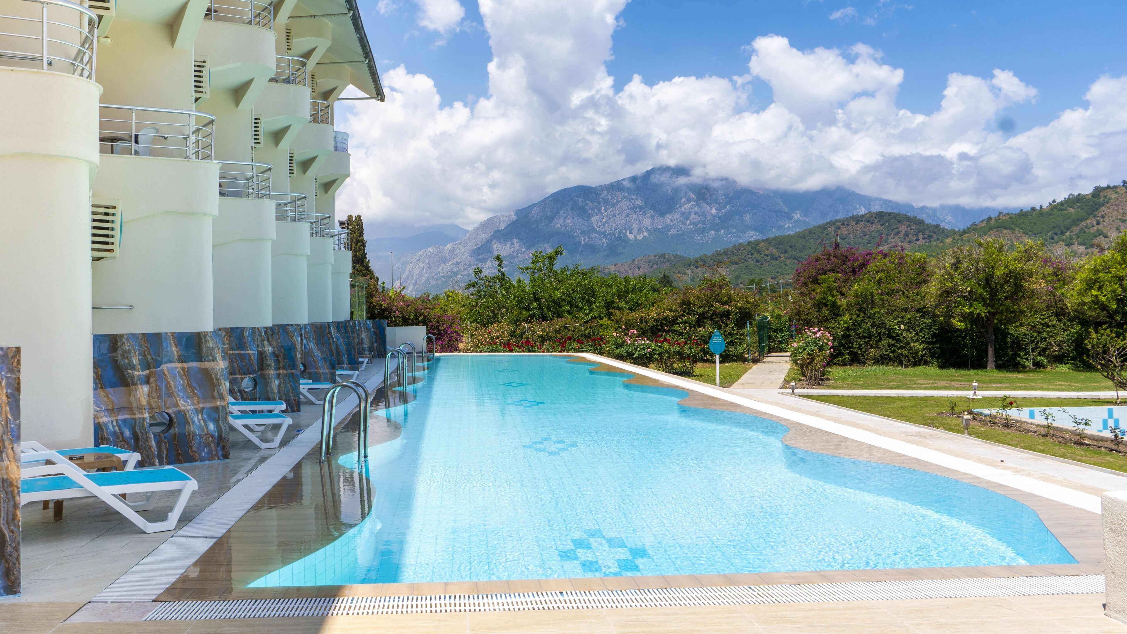 Una piscina con sillones de descanso, vistas a montañas y un edificio con balcones.