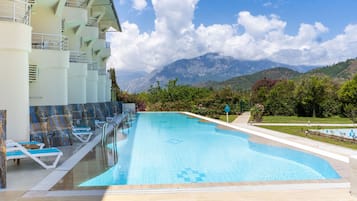 Una piscina con sillones de descanso, vistas a montañas y un edificio con balcones.
