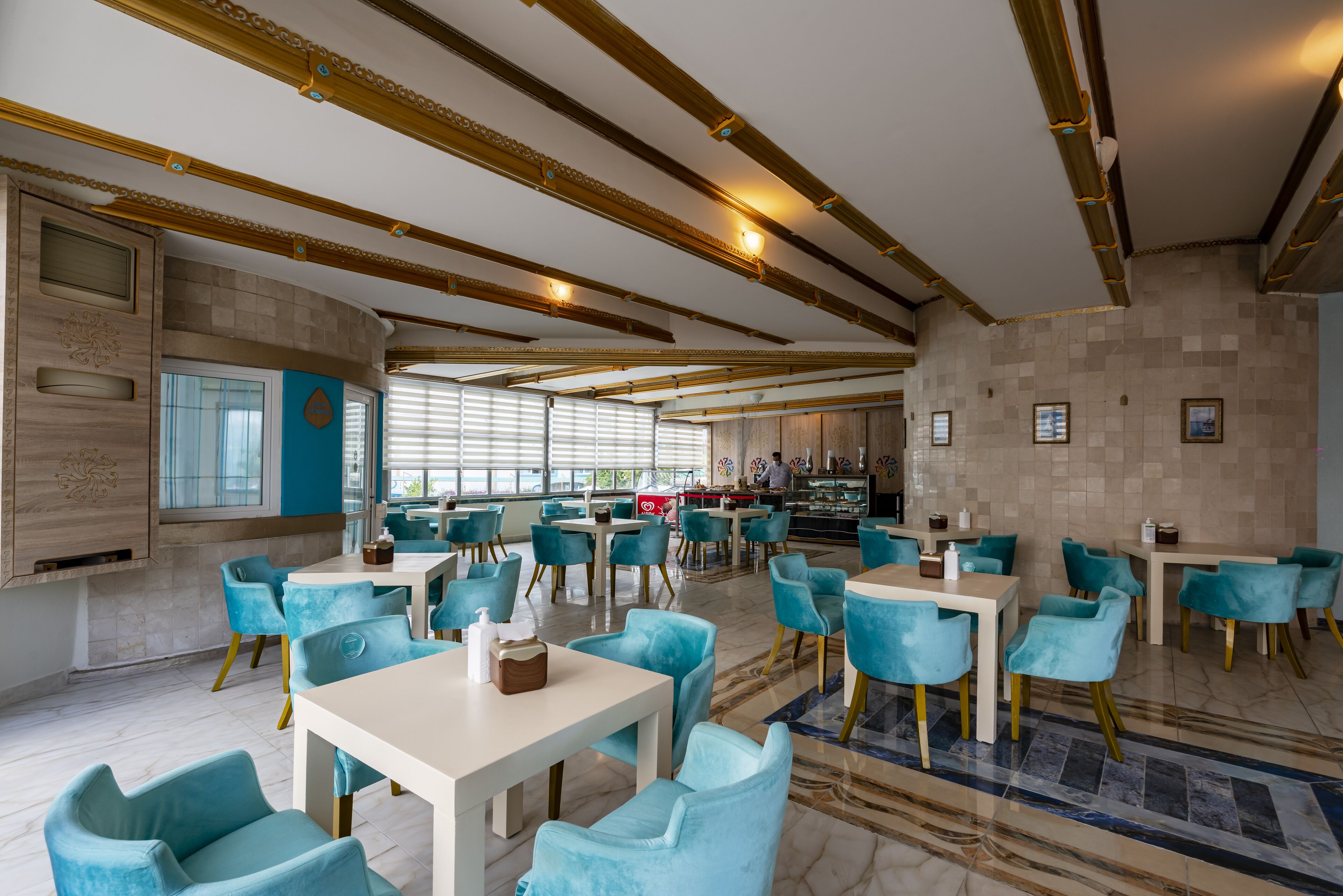A modern restaurant with teal chairs and white tables, featuring a marble floor and a high ceiling with exposed beams.