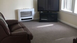 A living room with a brown sofa, a flat-screen TV on a black stand, a white heater, and a window with blinds. - Entire House 3 bedrooms, 5 minute walk to town centre and 1 minute to beach (Strahan)