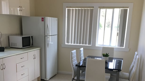 A kitchen with white cabinets, a microwave, and a refrigerator. There is a dining table with four chairs and a window with blinds. - Entire House 3 bedrooms, 5 minute walk to town centre and 1 minute to beach (Strahan)