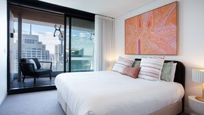 A modern hotel room with a large bed, a chair, a view of the city, and a colorful abstract painting. - Prestige City Chic I90 (Sydney)