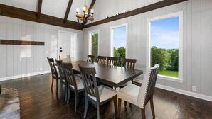 A dining room with a large wooden table and chairs, white paneled walls, and large windows offering a view of greenery. - Cabin on the Hill - North Nashville Stunner! (Hendersonville)
