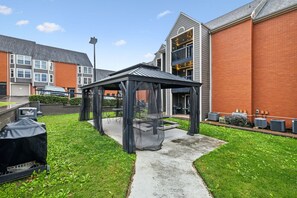 A covered outdoor seating area with a grill, surrounded by a brick building and a grassy lawn.