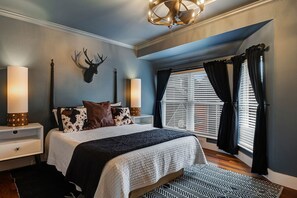 A bedroom with a bed, a mounted deer head, two bedside lamps, a window with curtains, and a patterned rug.