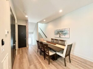A dining area with a long table and chairs, a staircase, and a painting on the wall.