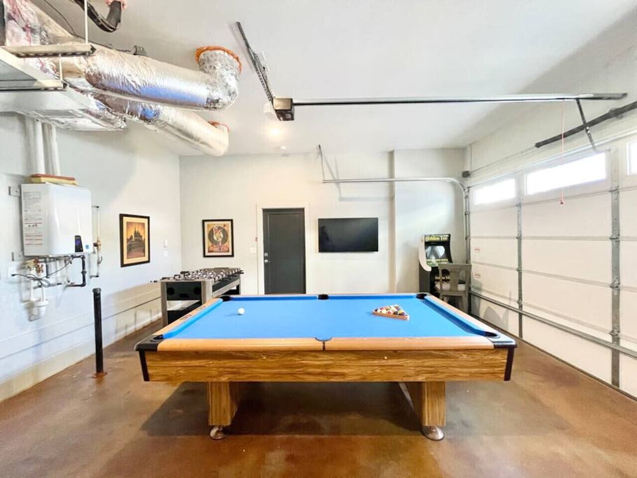 A pool table in a room with a garage door, a mounted TV, and a foosball table.