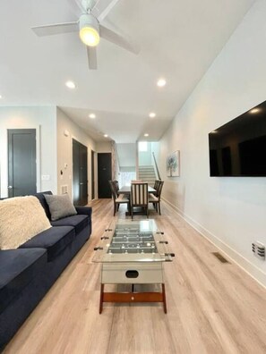 A modern living room with a sofa, a foosball table, a dining area with chairs, and a staircase.