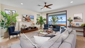 A spacious living room with a large window offering a view of the beach, a ceiling fan, and a comfortable seating arrangement. - Holua Kai #26: Luxury Fairway Front Villa w/ Pool, Spa, Lanai, AC & Ocean Views (Kailua-Kona)