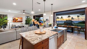 A modern kitchen with a large island, a dining area with a view of the ocean, and a living room with a TV and plants. - Holua Kai #26: Luxury Fairway Front Villa w/ Pool, Spa, Lanai, AC & Ocean Views (Kailua-Kona)
