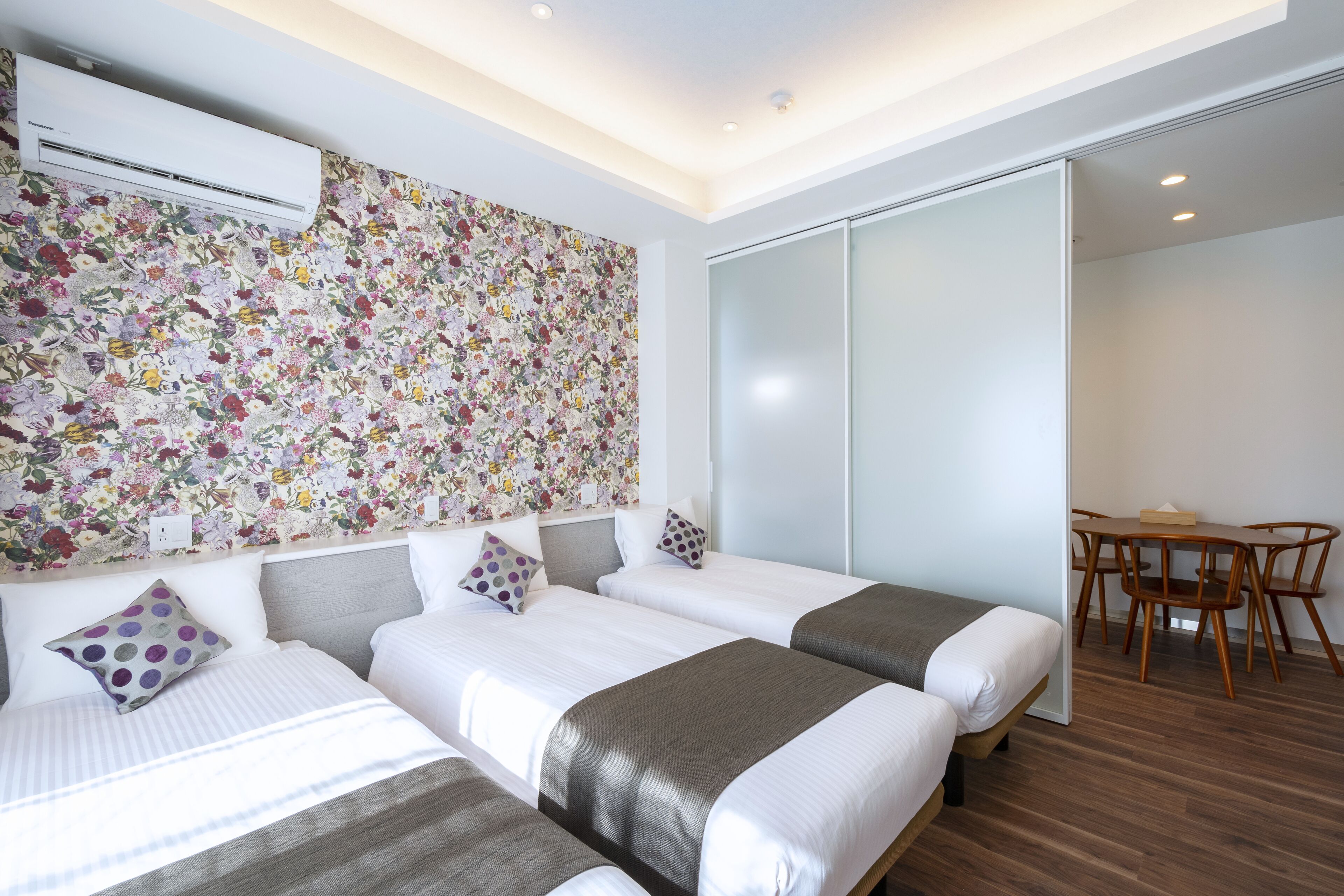 Une chambre d’hôtel avec trois lits, un mur décoré de motifs floraux, un climatiseur, une porte coulissante donnant sur un coin repas et un plancher en bois.