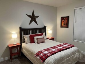 Uma cama bem arrumada com uma decoração de estrela acima, duas luminárias de cabeceira, um quadro na parede e uma janela com persianas.
