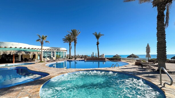 A resort pool area with multiple pools, palm trees, and a beach view.